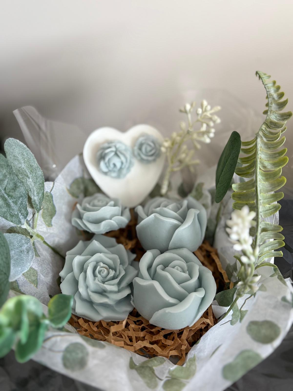 A decorative arrangement of four pale blue sculpted rose-shaped items, a small white heart-shaped ornament with two matching roses, and green foliage wrapped in white tissue paper on a neutral background.