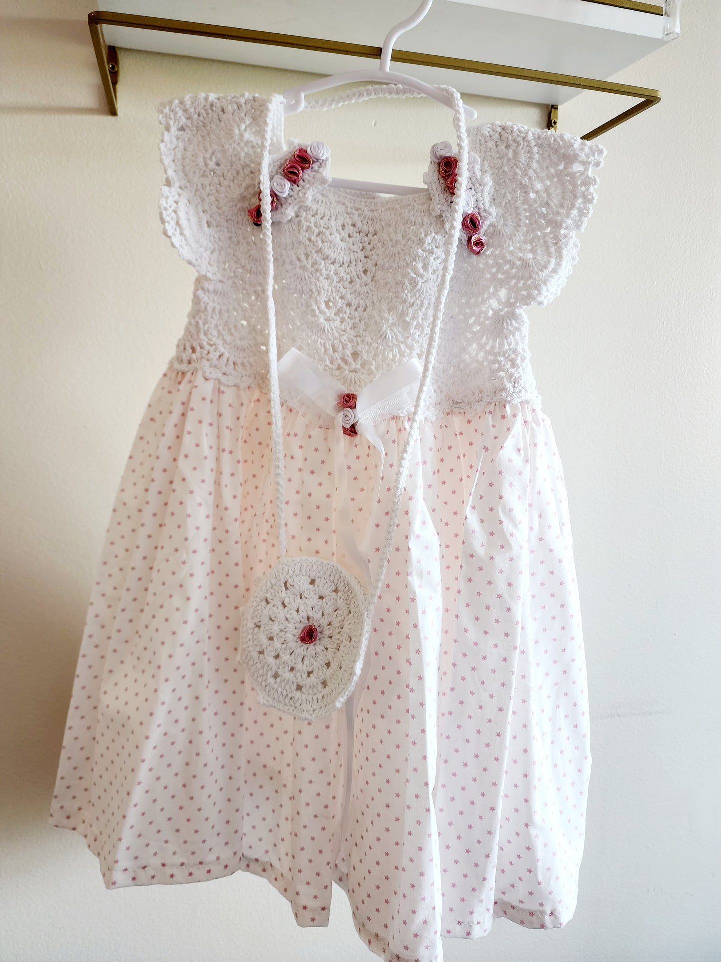 A white baby girl's dress with a crochet bodice, ruffled sleeves, and a white polka dot skirt, hanging on a white hanger with a matching crocheted crossbody bag.