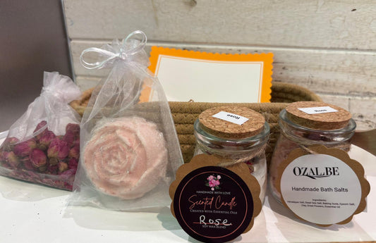 A spa gift set featuring a pink rose-shaped soap in a clear bag, a bag of dried pink rose petals, two glass jars of Ozalbe Handmade Bath Salts with cork lids, and a Scented Candle, arranged on a white surface with a woven basket and orange card in the background.