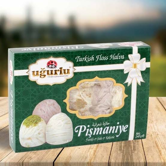A box of Ugurlu Turkish Floss Halva Pismaniye, featuring a dark green box with illustrations of three decorated halva balls and a window showing the creamy halva inside, set on a wooden surface with a blurred outdoor background.