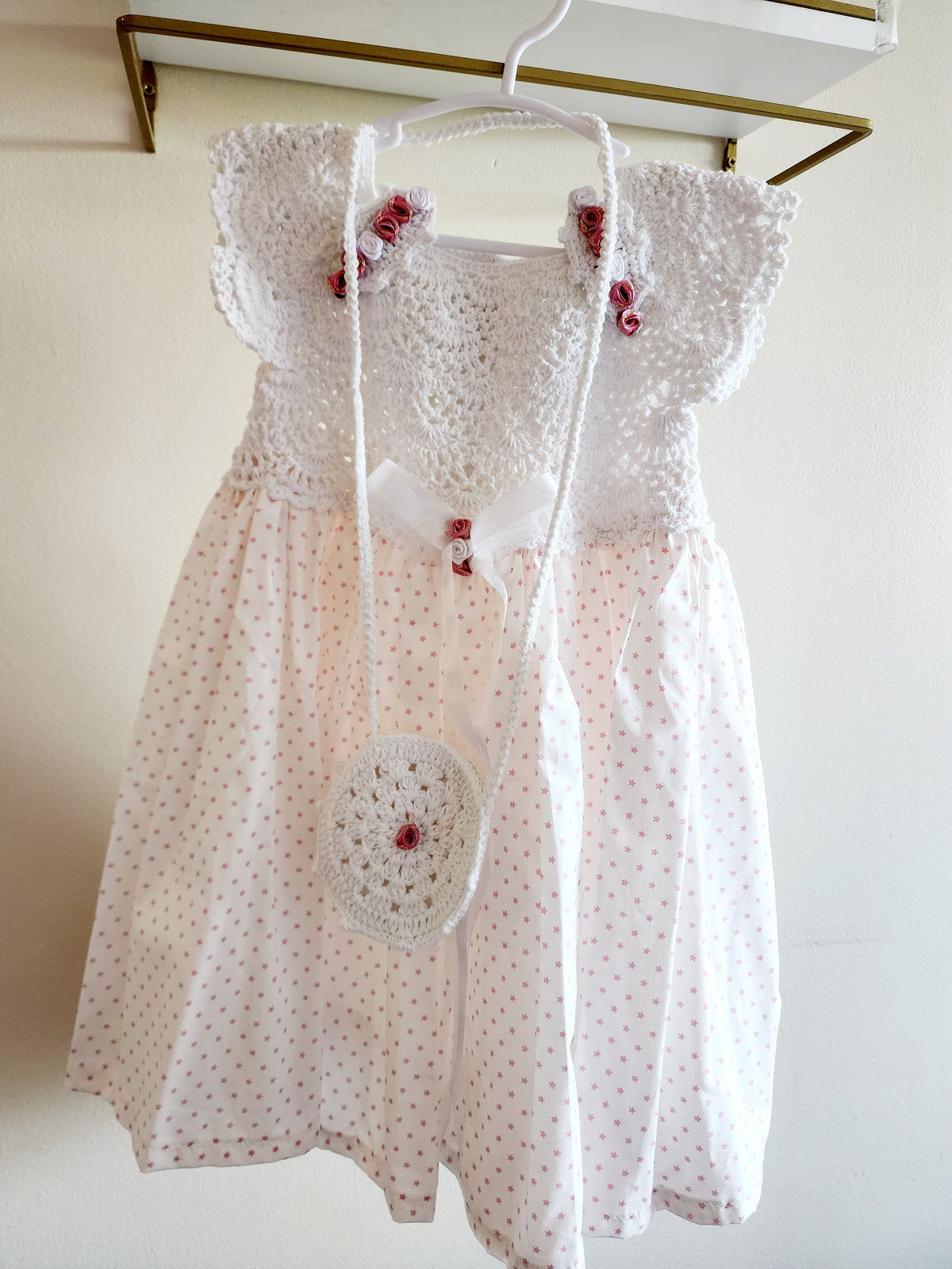 A white baby girl's dress with a crochet bodice, ruffled sleeves, and a white polka dot skirt, hanging on a white hanger with a matching crocheted crossbody bag.