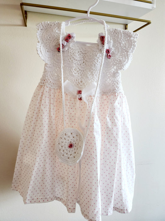A white baby girl's dress with a crochet bodice, ruffled sleeves, and a white polka dot skirt, hanging on a white hanger with a matching crocheted crossbody bag.