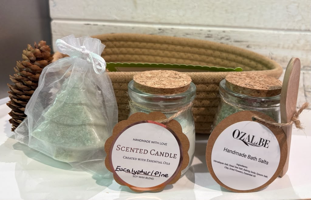 Two glass jars with cork lids labeled 'Scented Candle' and 'Handmade Bath Salts' sit on a white surface in front of a woven basket, alongside a clear bag of soap and a pinecone prop.