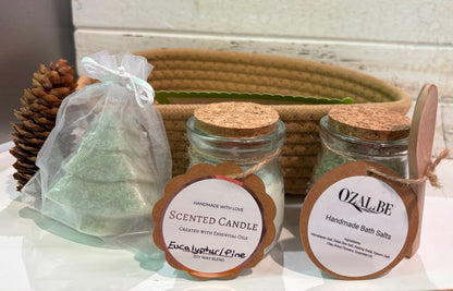 Two glass jars with cork lids labeled 'Scented Candle' and 'Handmade Bath Salts' sit on a white surface in front of a woven basket, alongside a clear bag of soap and a pinecone prop.