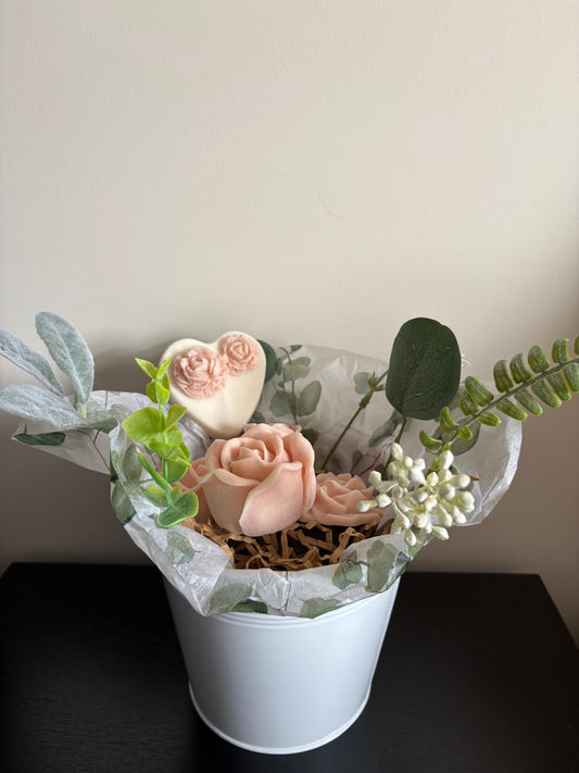 A floral arrangement in a white bucket with blush pink roses, green eucalyptus leaves, white berries, and a heart-shaped soap or wax flower, set against a plain beige wall.