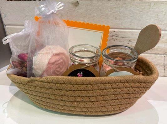 A spa gift basket set in a woven natural  basket, containing soap, bath salt, candle, and a bag of dried flowers, set against a rustic white wooden background.