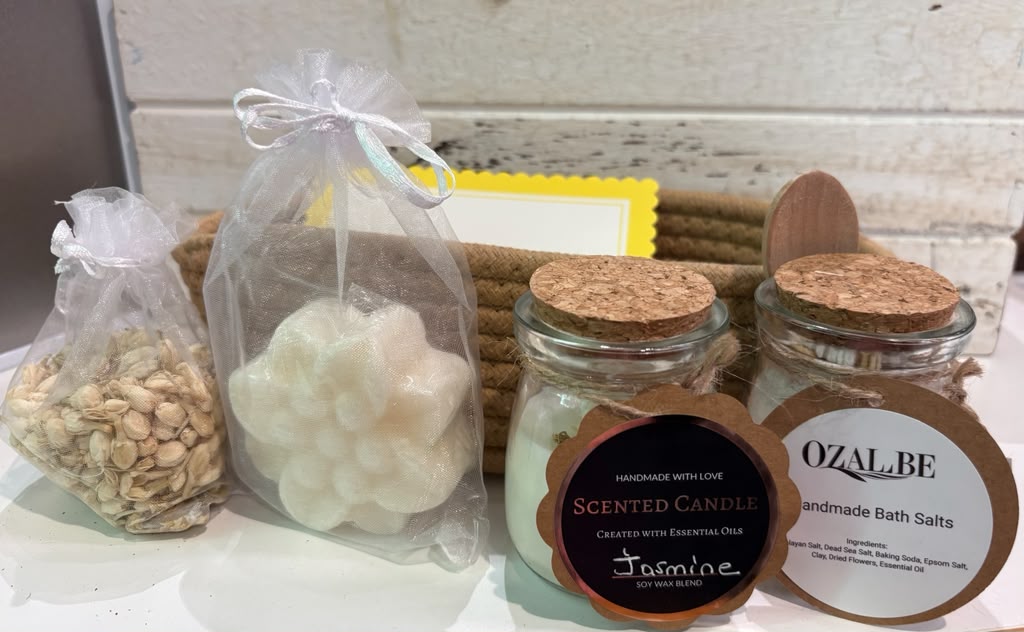 A collection of handmade bath and body products including a white flower-shaped candle in a clear bag, a bag of bath salts, and two glass jars of scented candle and ozalbe handmade bath salts with cork lids.