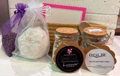A bath gift set featuring a lavender soap in a sheer purple drawstring bag, two glass jars of handmade candle and bath salts with cork lids, and two labels reading 'Scented Oils Lavender' and 'OZALBE Handmade Bath Salts'.