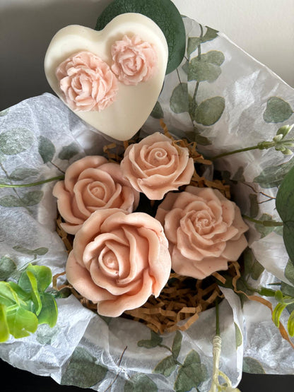 A decorative gift arrangement featuring four pale blush pink rose-shaped items in a basket with shredded brown paper, surrounded by white tissue paper printed with green leaves, a white heart-shaped item with pink rose details, and green leaves.