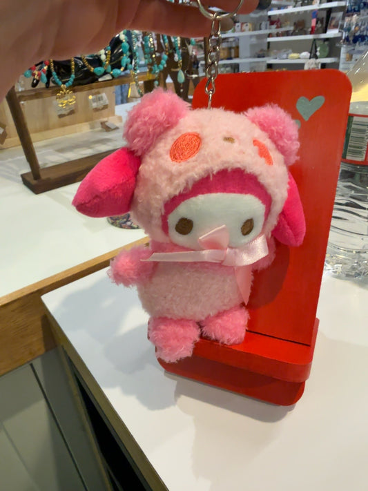 A pink plush keychain character with a white face and pink ears, hanging from a metal ring in a retail store setting, positioned in front of a red display stand.