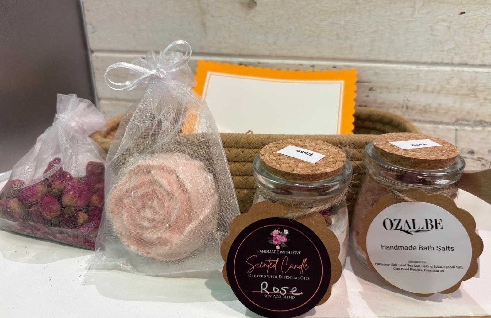 A spa gift set featuring a pink rose-shaped soap in a clear bag, a bag of dried pink rose petals, two glass jars of Ozalbe Handmade Bath Salts with cork lids, and a Scented Candle, arranged on a white surface with a woven basket and orange card in the background.
