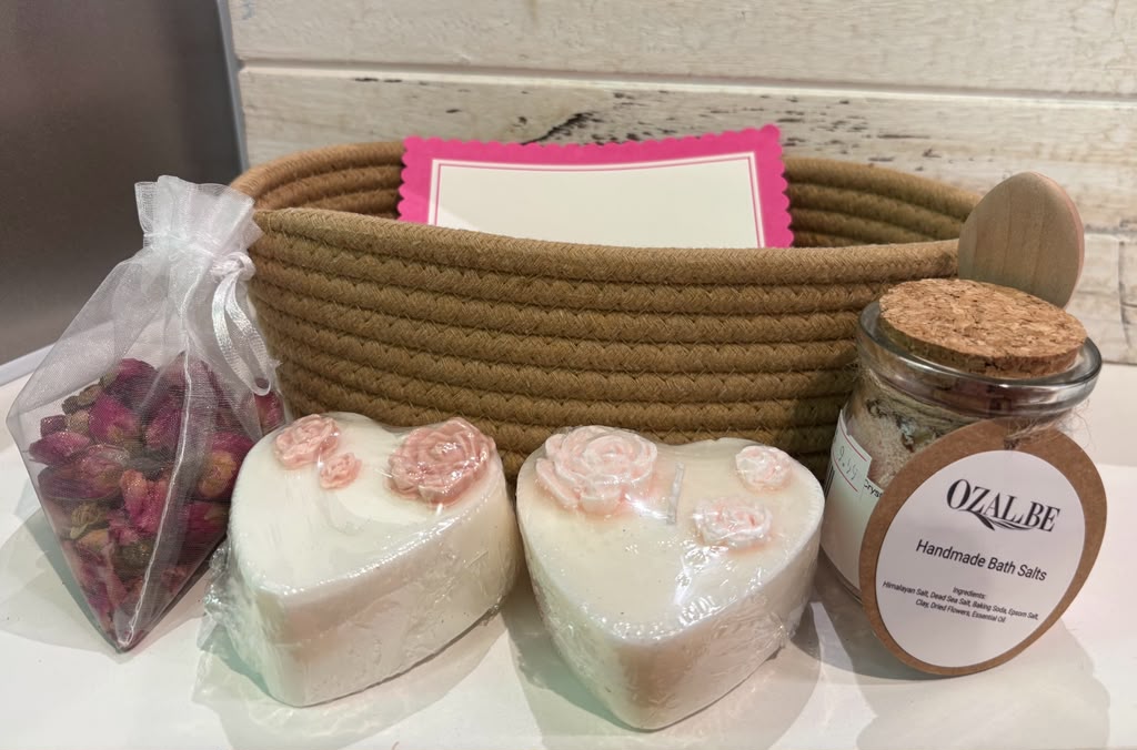 Two heart-shaped handmade soap and candle with pink rose decorations, a jar of OZALBE Handmade Bath Salts, a bag of dried rose petals, and a brown rope basket with a pink card, arranged on a white surface.