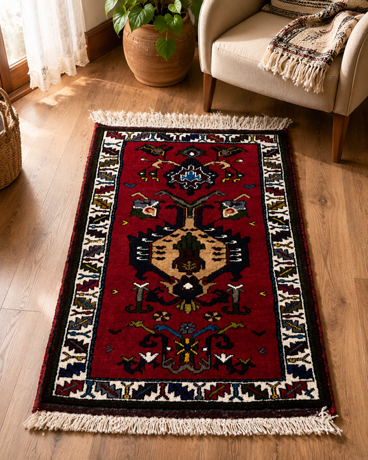 A rectangular red area rug with a traditional geometric pattern and cream fringe, shown on a light wood floor in a living room setting next to a beige armchair and a potted plant.