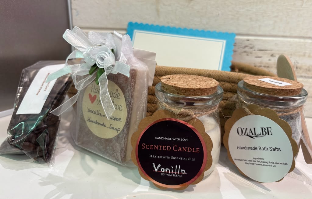 A gift set containing a scented vanilla candle in a glass jar, a glass jars of handmade bath salts, vanilla coffee soap and a bag of organic coffee. Set against a rustic white wooden background.