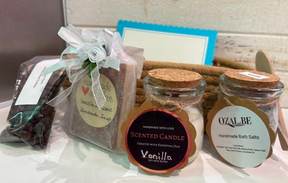 A gift set containing a scented vanilla candle in a glass jar, a glass jars of handmade bath salts, vanilla coffee soap and a bag of organic coffee. Set against a rustic white wooden background.